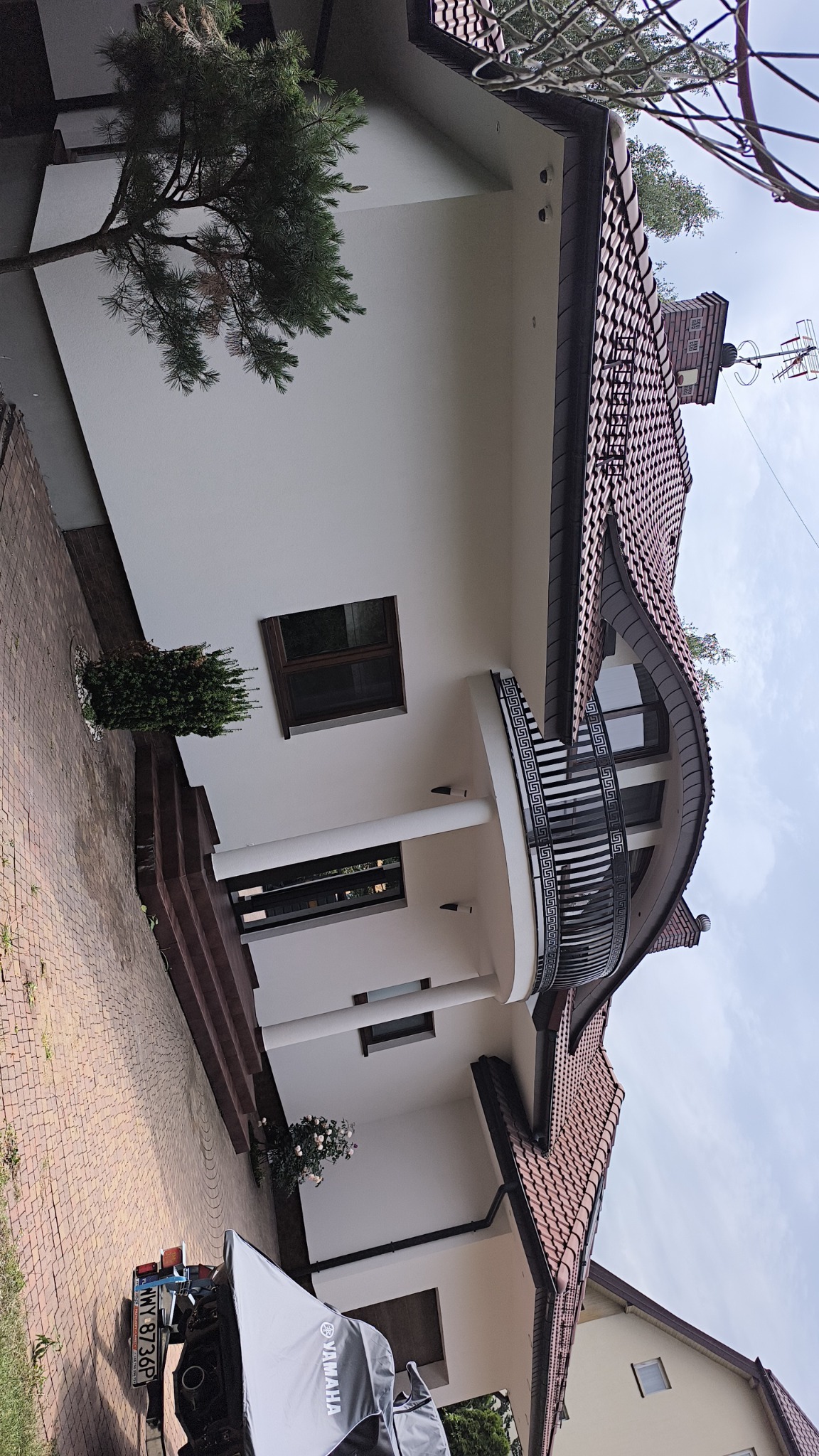 Nowoczesny dom jednorodzinny z białymi ścianami, brązowym dachem z dachówki i balkonem z ozdobną balustradą, widoczny z poziomu chodnika wyłożonego kostką.