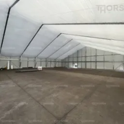 Wnętrze dużej hali z metalową konstrukcją szkieletową i białym, napiętym poszyciem dachowym, widoczny podnośnik nożycowy w lewym rogu.