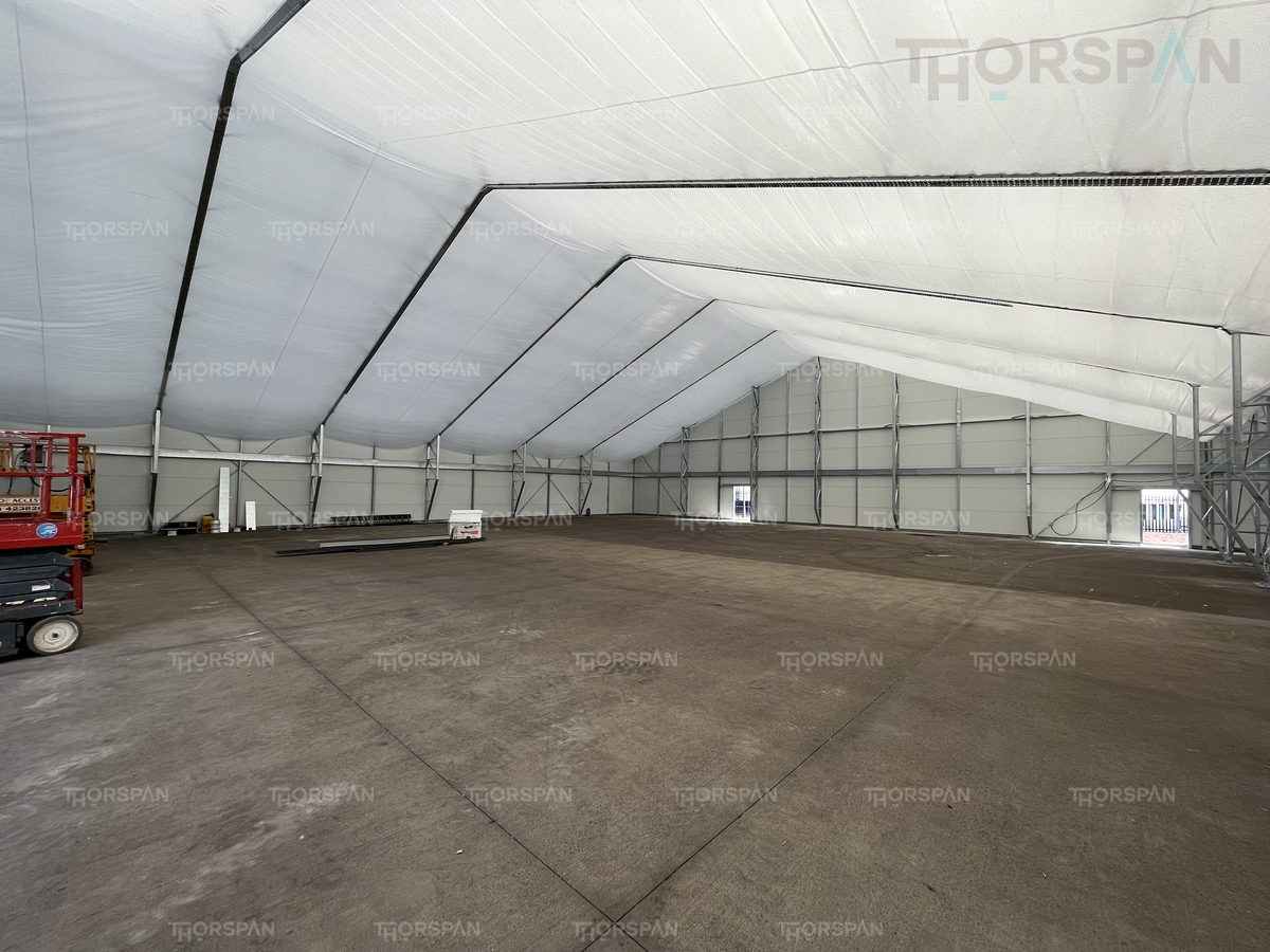 Wnętrze dużej hali z metalową konstrukcją szkieletową i białym, napiętym poszyciem dachowym, widoczny podnośnik nożycowy w lewym rogu.