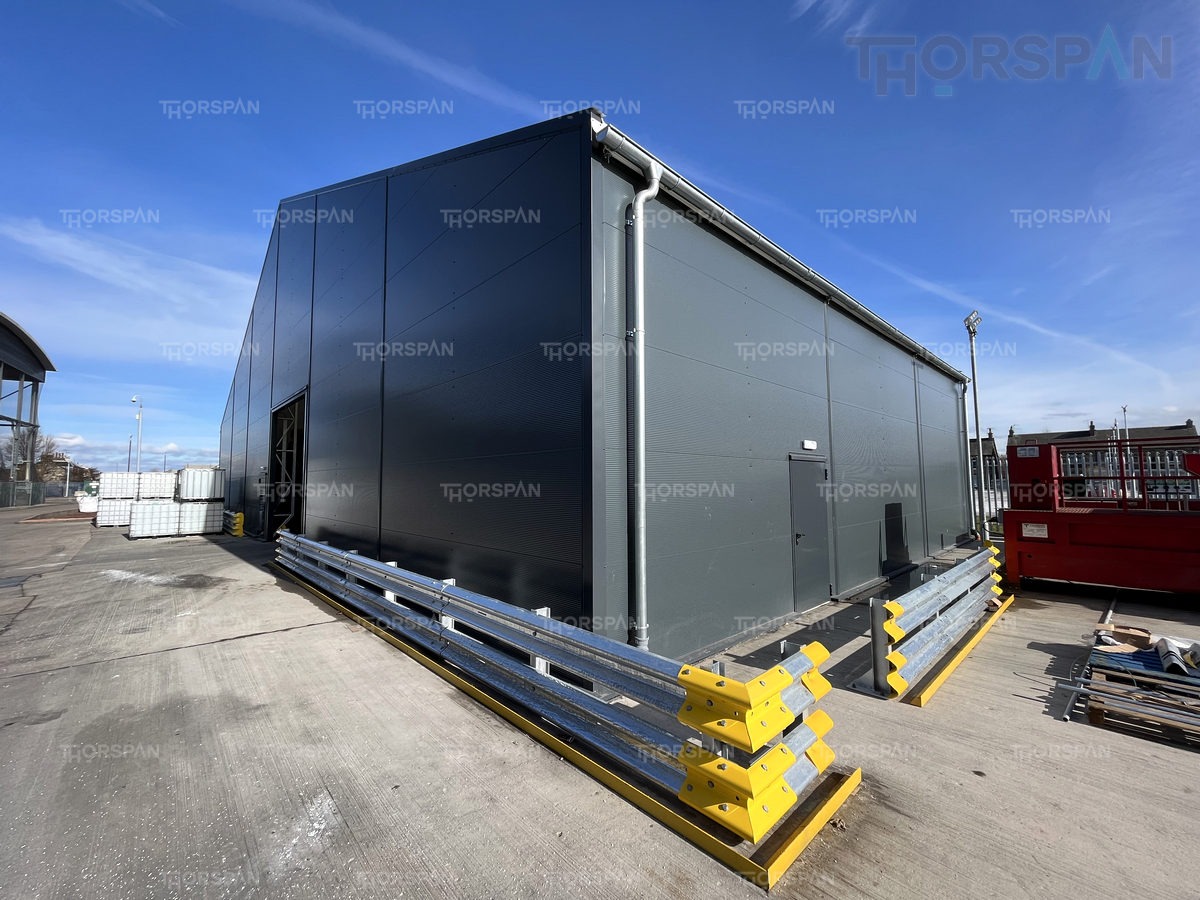 Gotowa, szara hala stalowa z widocznym systemem rynnowym i zabezpieczeniami w postaci barier ochronnych z żółtymi elementami, na tle błękitnego nieba.