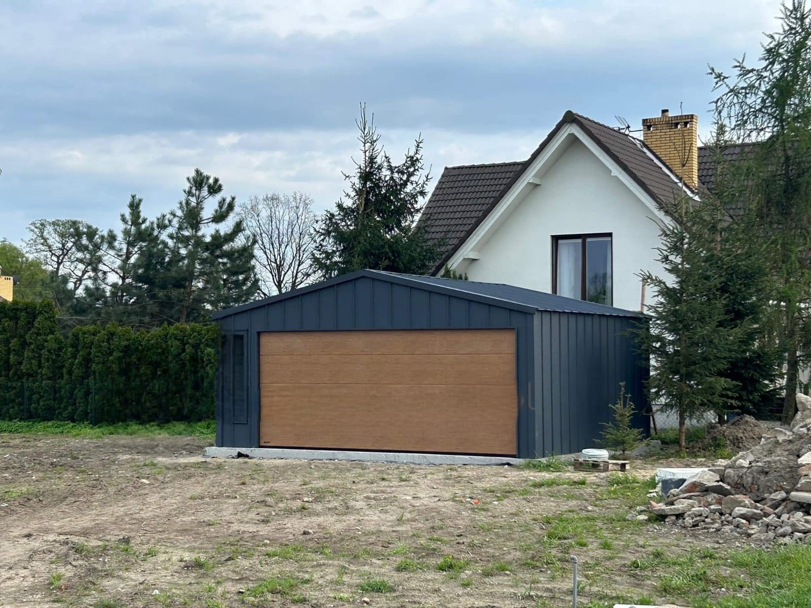 Nowoczesny garaż blaszany z segmentową bramą w kolorze drewna, na tle domu jednorodzinnego i zieleni, widoczny niezagospodarowany teren.