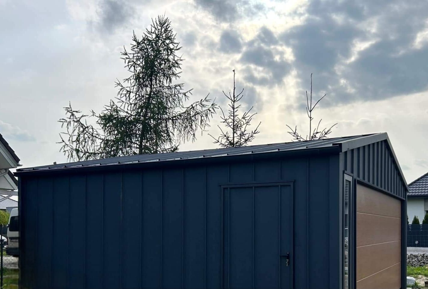 Nowoczesny, ciemnoniebieski budynek gospodarczy z płaskim dachem i brązowymi akcentami, na tle pochmurnego nieba i drzew iglastych.