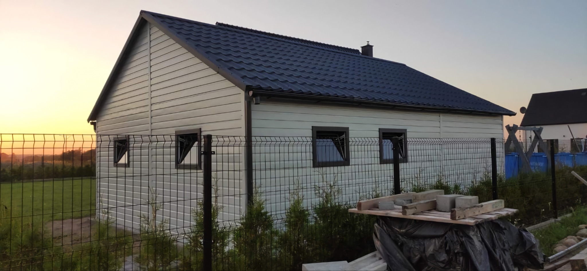 Gotowy garaż z blachy trapezowej, obłożony sidingiem, z trzema oknami, ogrodzony siatką, na tle zachodzącego słońca.