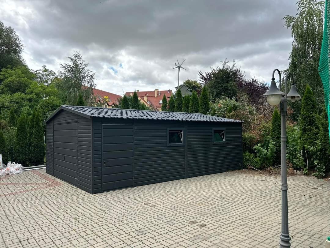 Nowoczesny, antracytowy garaż z blachy trapezowej z dwuspadowym dachem, oświetlony latarnią uliczną, na tle zieleni i pochmurnego nieba.