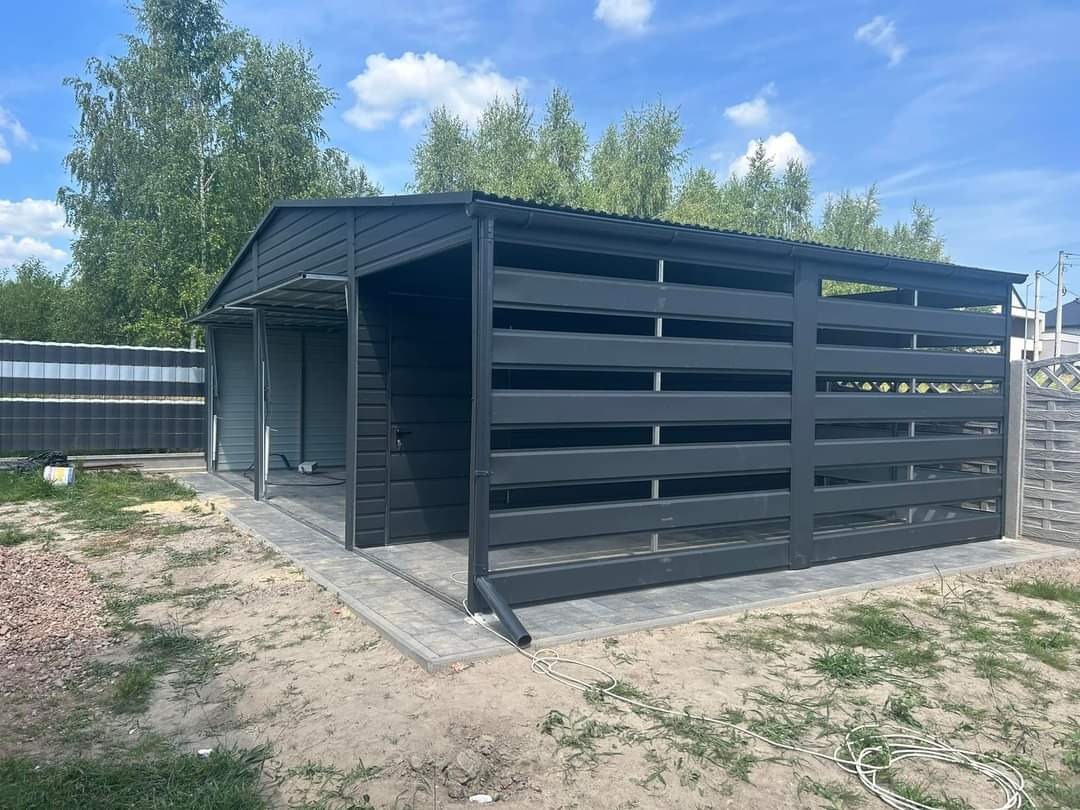 Nowoczesna, ciemnoszara konstrukcja z metalu, prawdopodobnie garaż lub wiata, z poziomymi panelami i zadaszeniem, ustawiona na kostce brukowej, z widocznym przewodem elektrycznym na ziemi.