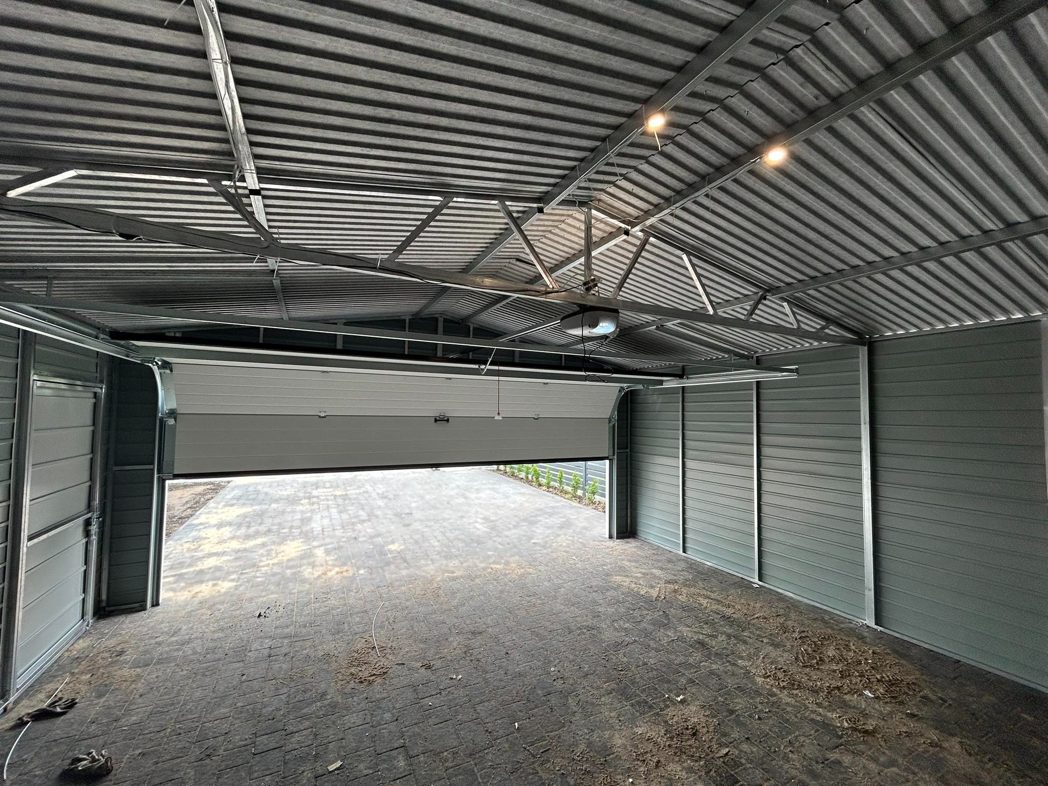 Wnętrze garażu blaszanego z częściowo otwartą bramą segmentową w Bratysławie, widoczny mechanizm automatycznego otwierania bramy pod sufitem i posadzka z kostki brukowej.