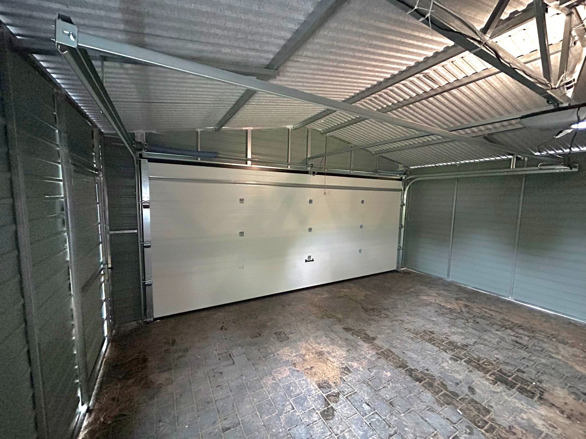 Wnętrze garażu blaszanego z częściowo otwartą białą segmentową bramą garażową, widoczna metalowa konstrukcja dachu i ściany z blachy trapezowej.