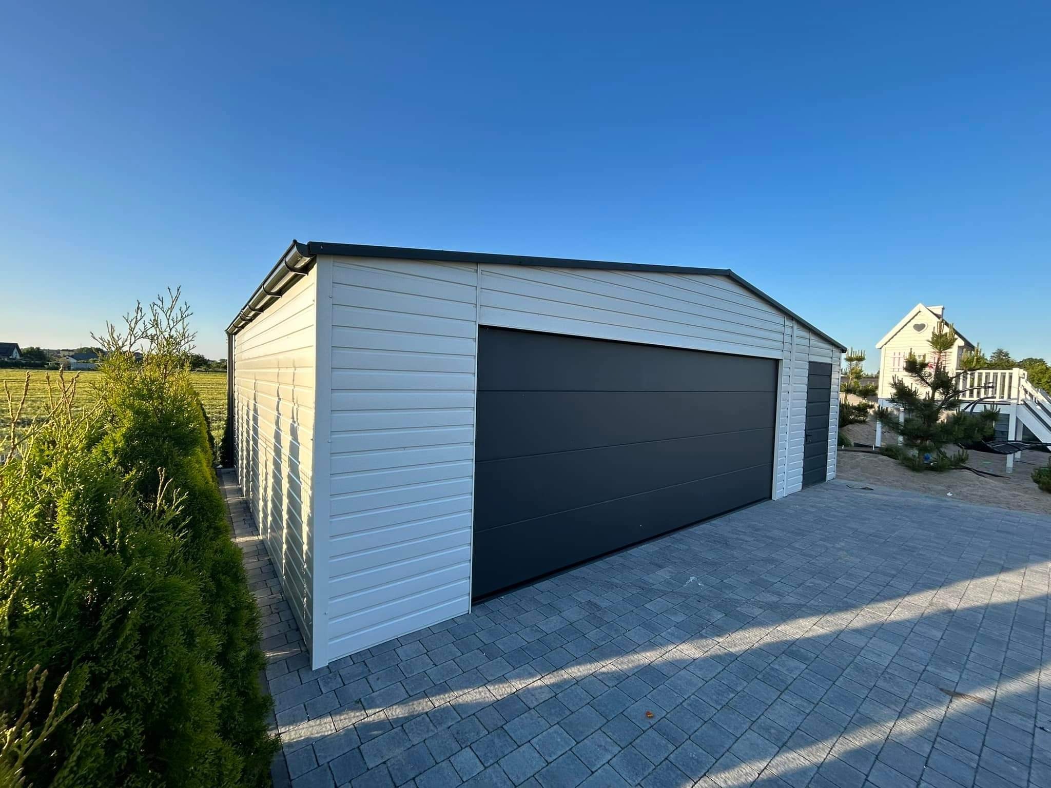 Nowoczesny, biało-szary garaż z paneli, z ciemną bramą segmentową, na posesji z kostką brukową i tujami, w słoneczny dzień.