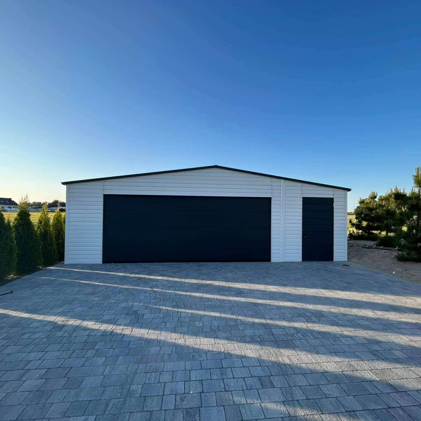 Nowoczesny, biały garaż z czarną bramą segmentową i bocznymi drzwiami, wykończony na zewnątrz panelami, położony na brukowanym podjeździe w słoneczny dzień.