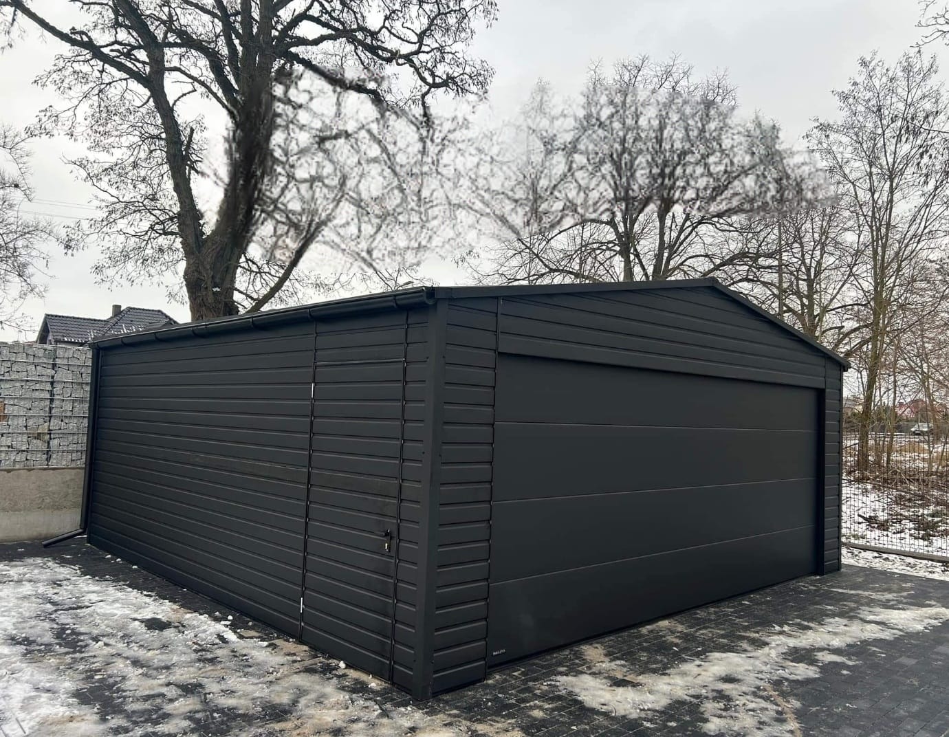 Czarny garaż blaszany z bramą segmentową, widoczny na tle drzew i kostki brukowej z resztkami śniegu.