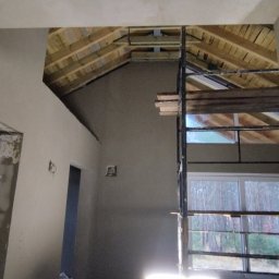 Tynk tynk - Wnętrze w trakcie prac wykończeniowych: ściany pokryte świeżym tynkiem, widoczne drewniane elementy konstrukcji dachu oraz rusztowanie przed oknem z widokiem na las.