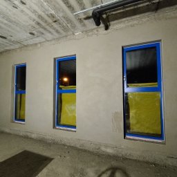 Tynk tynk - Ściana po nałożeniu surowego tynku cementowo-wapiennego z widocznymi trzema oknami z niebieskimi ramami, zabezpieczonymi folią. Widoczny betonowy sufit z instalacjami.