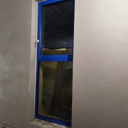 Tynk tynk - Świeżo nałożony tynk wokół niebieskiej ramy okiennej, widoczne nierówności i szorstka faktura przed dalszą obróbką.