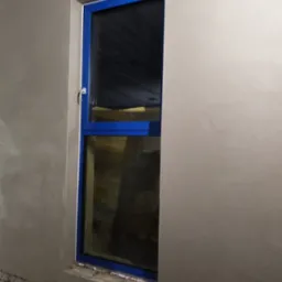 Świeżo nałożony tynk wokół niebieskiej ramy okiennej, widoczne nierówności i szorstka faktura przed dalszą obróbką.