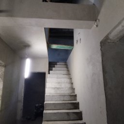 Tynk tynk - Surowe schody betonowe w trakcie budowy lub remontu, widoczne ściany w stanie surowym, brak wykończenia, prowizoryczne oświetlenie, fragment instalacji elektrycznej na ścianie.