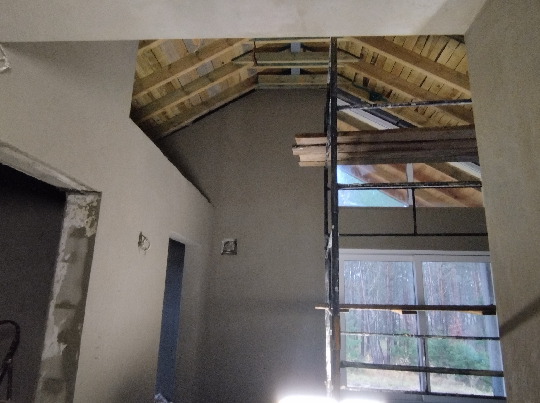 Wnętrze w trakcie prac wykończeniowych: ściany pokryte świeżym tynkiem, widoczne drewniane elementy konstrukcji dachu oraz rusztowanie przed oknem z widokiem na las.