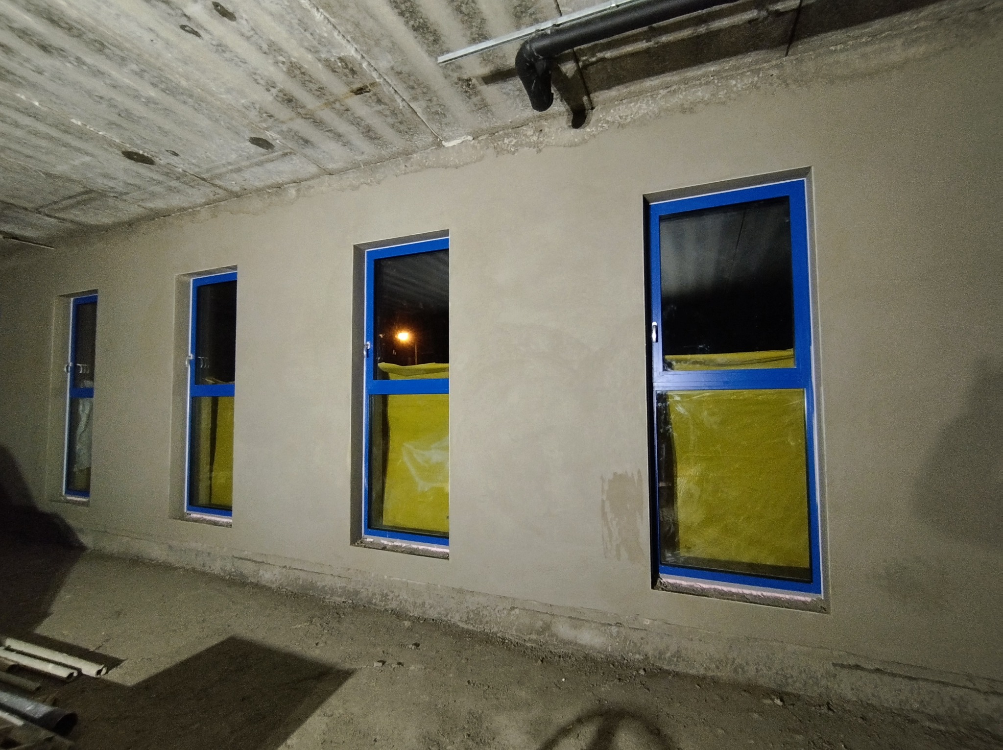 Ściana po nałożeniu surowego tynku cementowo-wapiennego z widocznymi trzema oknami z niebieskimi ramami, zabezpieczonymi folią. Widoczny betonowy sufit z instalacjami.