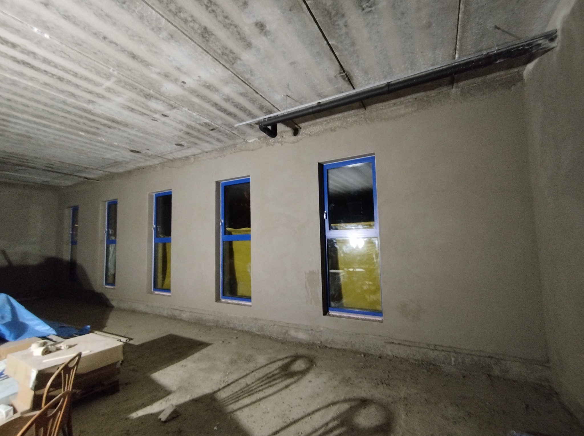 Wnętrze surowego budynku z nowymi oknami z niebieskimi ramami, ściany pokryte świeżo nałożoną warstwą szarego tynku, widoczny surowy betonowy strop i instalacje pod sufitem.