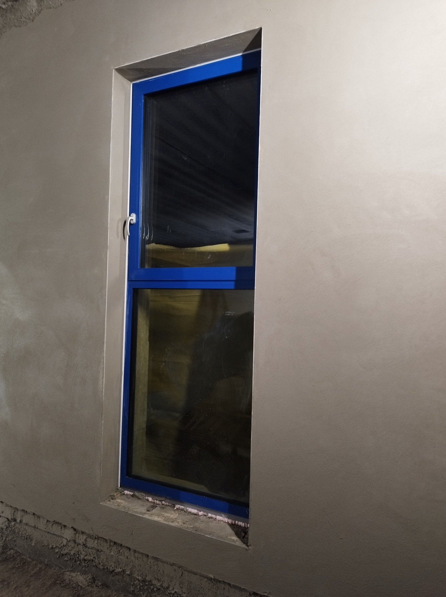 Świeżo nałożony tynk wokół niebieskiej ramy okiennej, widoczne nierówności i szorstka faktura przed dalszą obróbką.