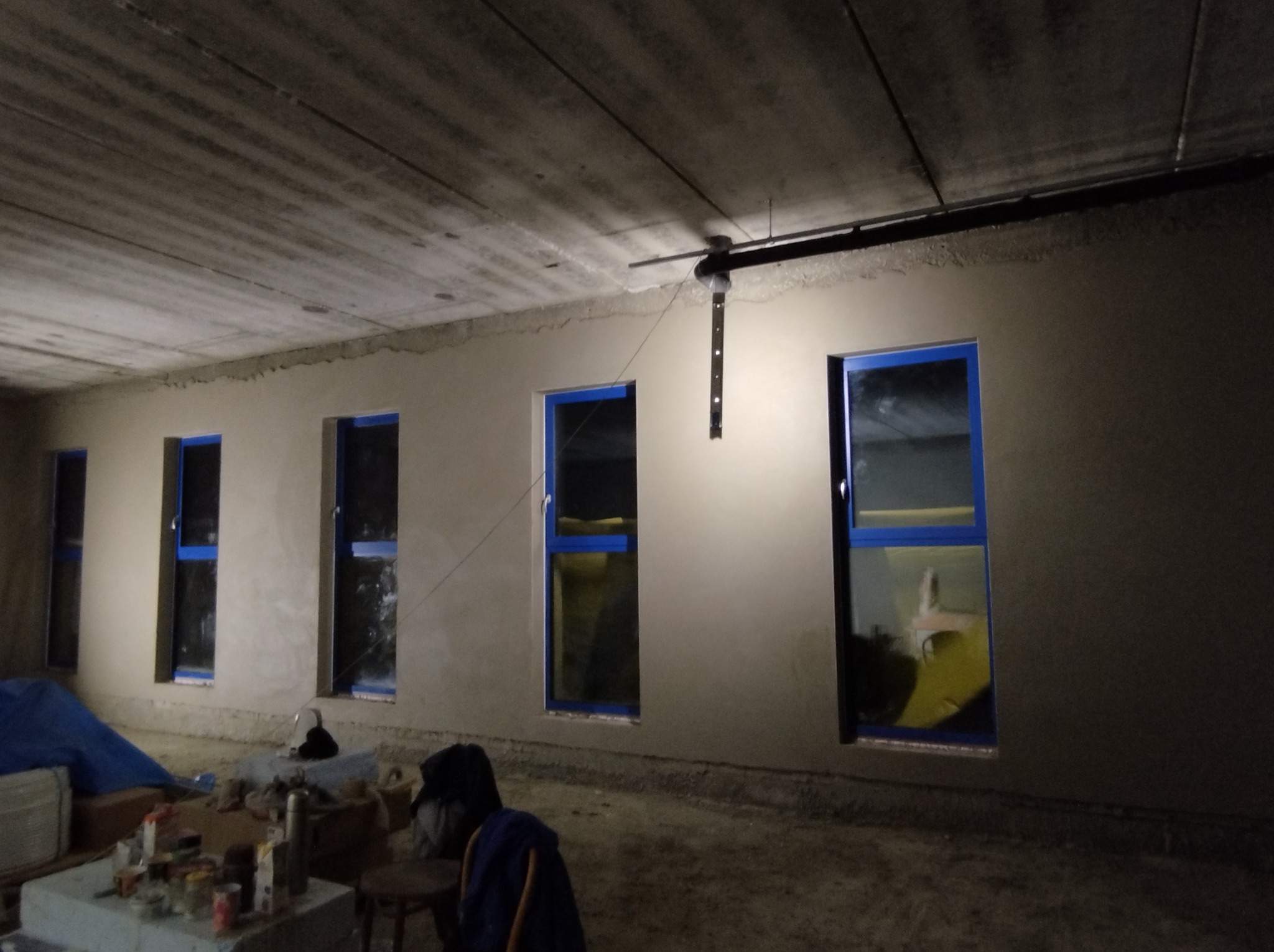 Wnętrze w trakcie prac wykończeniowych: ściany z surowym tynkiem, okna z niebieską taśmą zabezpieczającą, widoczne narzędzia i materiały budowlane na podłodze.