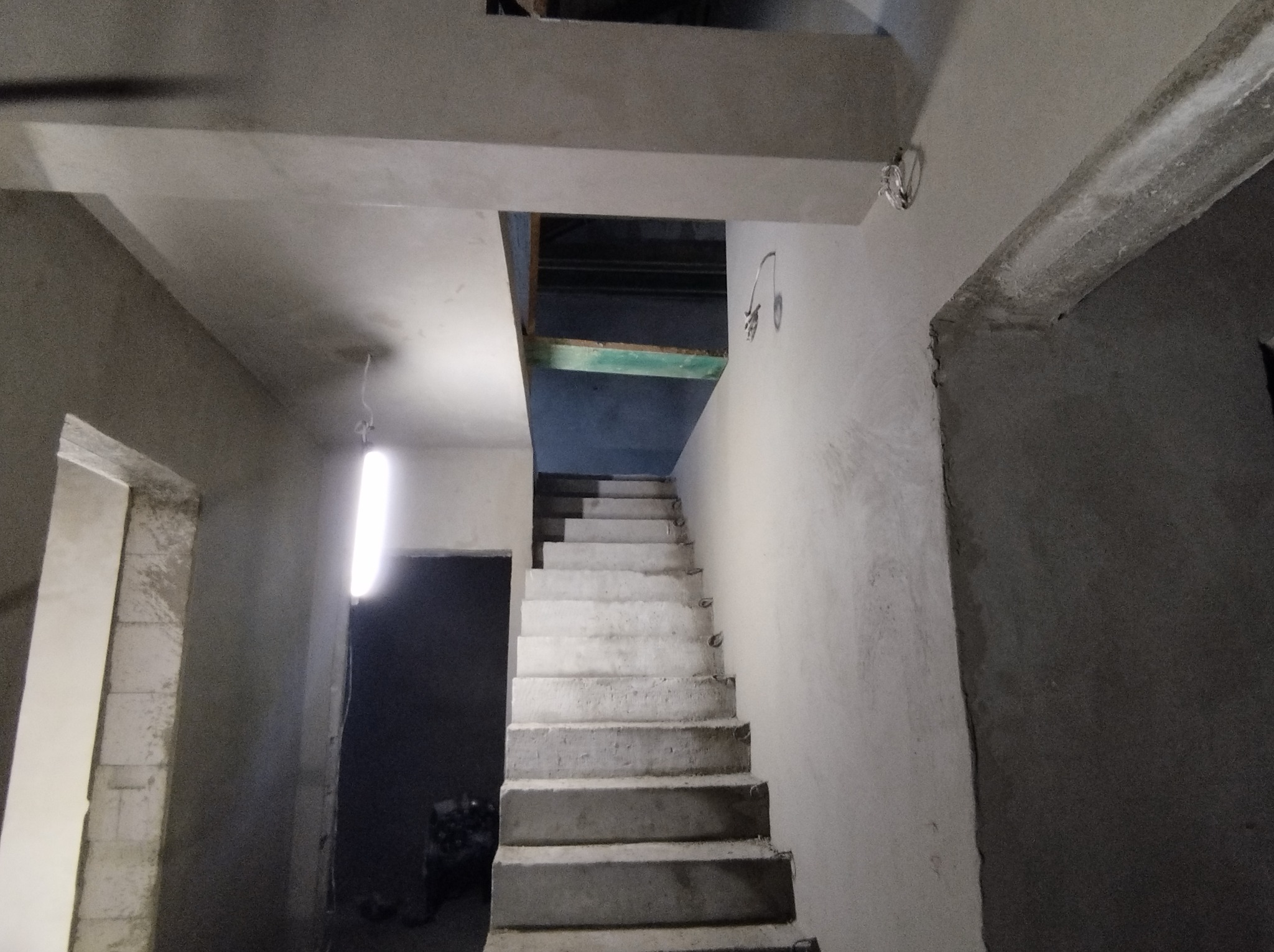 Surowe schody betonowe w trakcie budowy lub remontu, widoczne ściany w stanie surowym, brak wykończenia, prowizoryczne oświetlenie, fragment instalacji elektrycznej na ścianie.