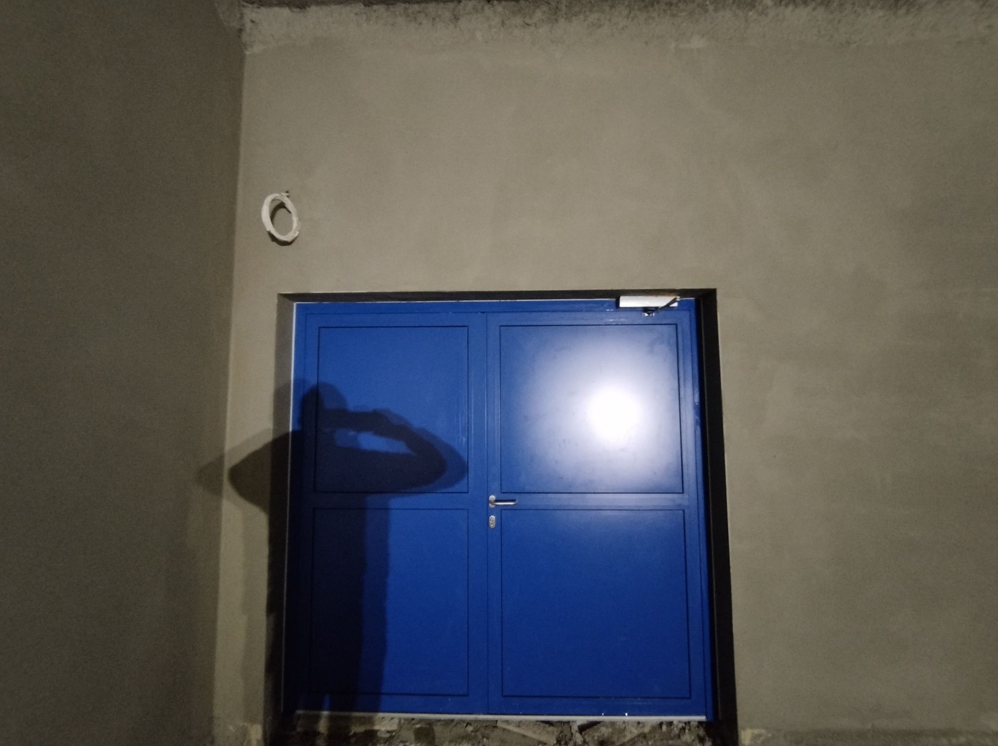 Świeżo położone tynki na ścianie wewnętrznej, widoczna niebieska, metalowa brama garażowa z ciemnym cieniem osoby. Ściana szara, surowa, z widocznymi niedoskonałościami przy suficie.
