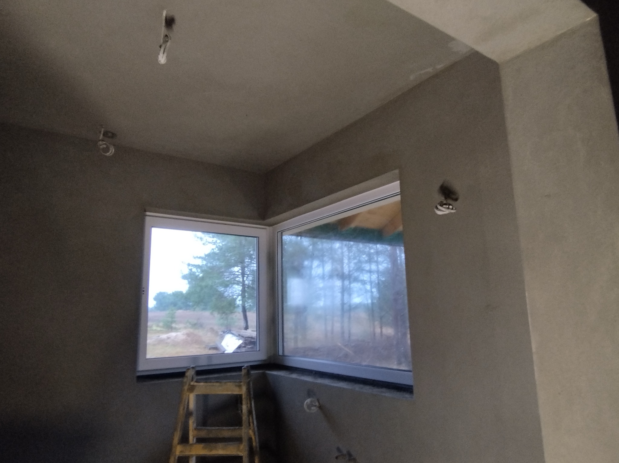 Pokój w stanie deweloperskim z surowymi ścianami i dwoma oknami narożnymi, przez które widać las; widoczna drabina malarska.