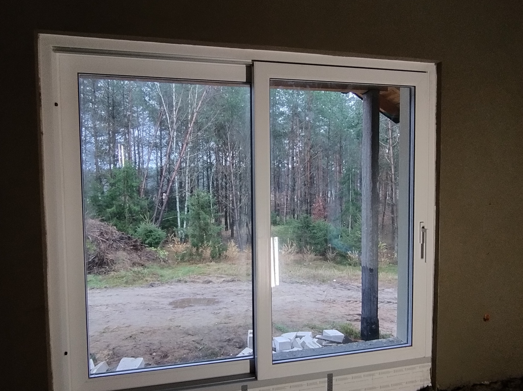 Wnętrze z zamontowanym przesuwnym oknem PCV z widokiem na zalesiony teren. Ściana w surowym stanie przed wykończeniem.