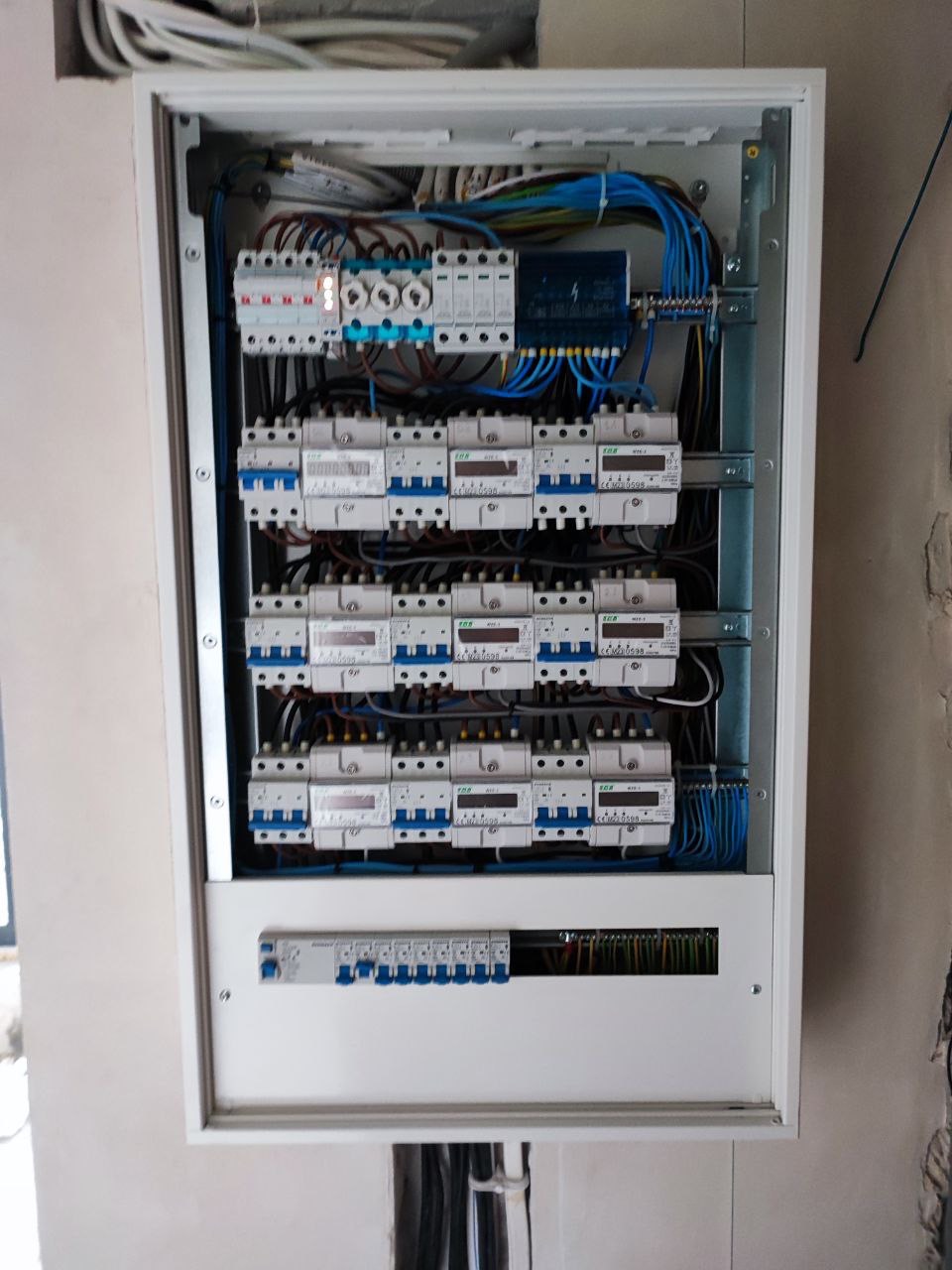 Rozdzielnica elektryczna w białej obudowie, widoczne liczne bezpieczniki, kable i przewody, częściowo zamontowana w ścianie w trakcie prac wykończeniowych.