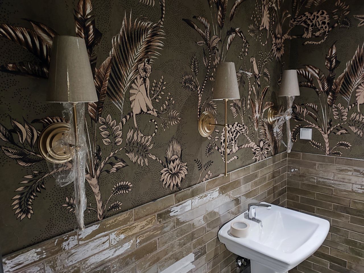 Ścienne lampy z abażurami, jedna owinięta folią ochronną, zamontowane na ścianie w łazience z tapetą w motywy roślinne i zwierzęce; umywalka z baterią w dolnej części kadru.