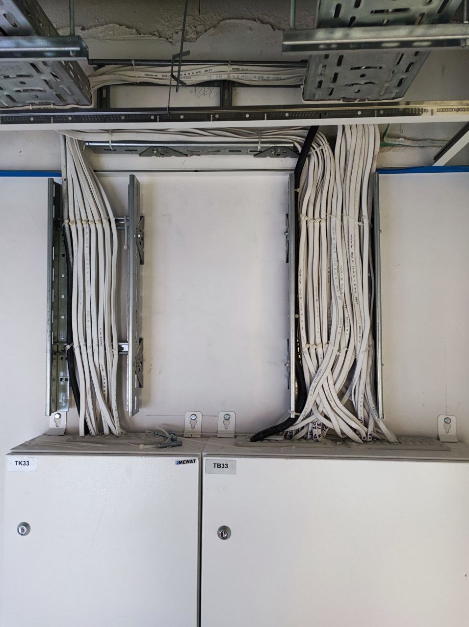 Białe kable elektryczne prowadzone w metalowych korytach pod sufitem, wchodzące do szarej skrzynki elektrycznej oznaczonej TK33 i TB33.