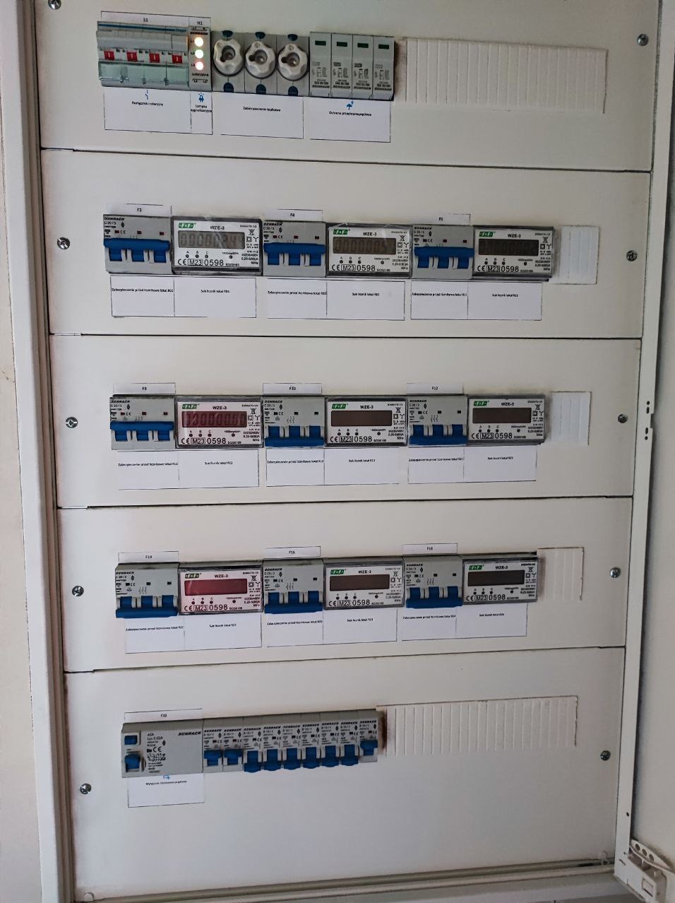 Otwarta skrzynka elektryczna z zainstalowanymi licznikami energii i bezpiecznikami, widoczne liczniki z wyświetlaczami i oznaczeniami producenta, biała obudowa skrzynki.