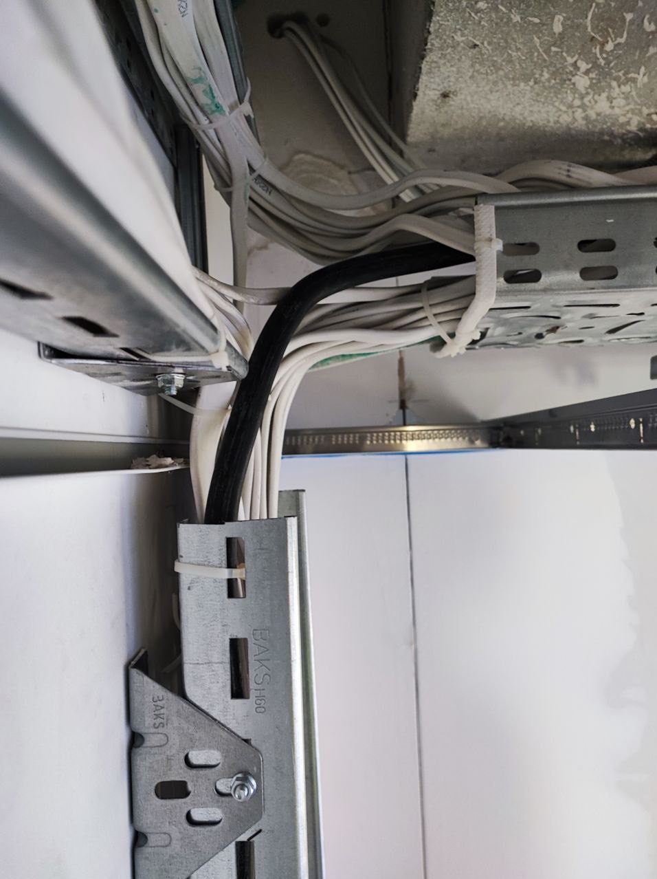 Przewody elektryczne prowadzone w korycie kablowym, mocowane do sufitu, widoczne białe i czarne kable spięte opaskami zaciskowymi.