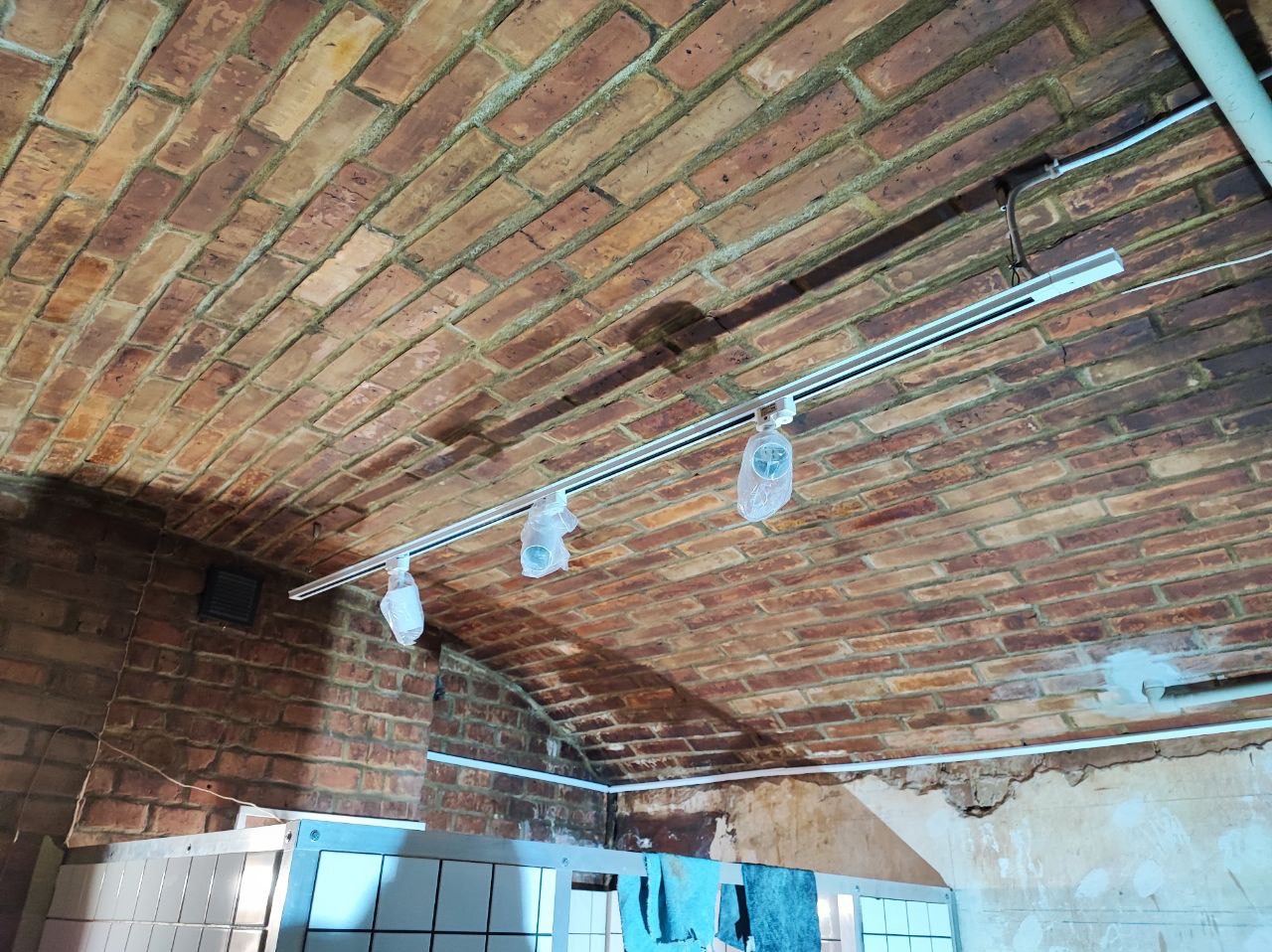 Szyna oświetleniowa zamontowana na ceglanym sklepieniu łukowym, z trzema lampami w foliowych osłonach ochronnych, widoczne okablowanie i fragment pomieszczenia z białymi płytkami.