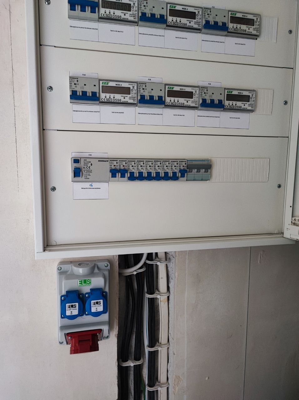 Rozdzielnica elektryczna z zabezpieczeniami i licznikami, widoczne przewody w korytku ściennym oraz podwójne gniazdo elektryczne ELS z czerwonym wyłącznikiem.