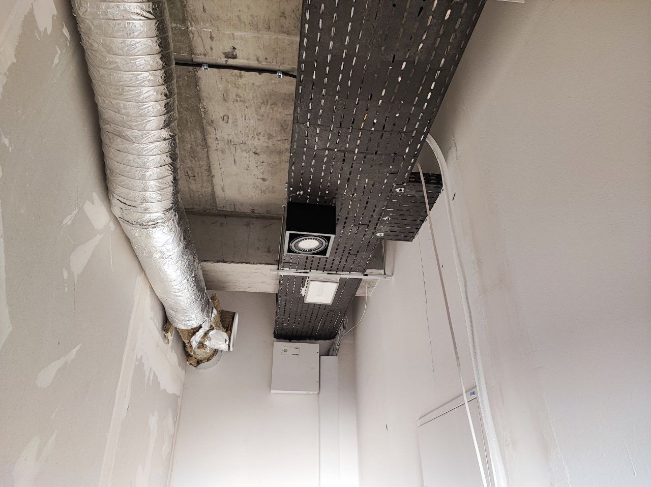 Szara, perforowana korytka kablowa pod sufitem z zamontowanym oświetleniem punktowym LED i rurą wentylacyjną w surowym stanie wykończenia wnętrza.