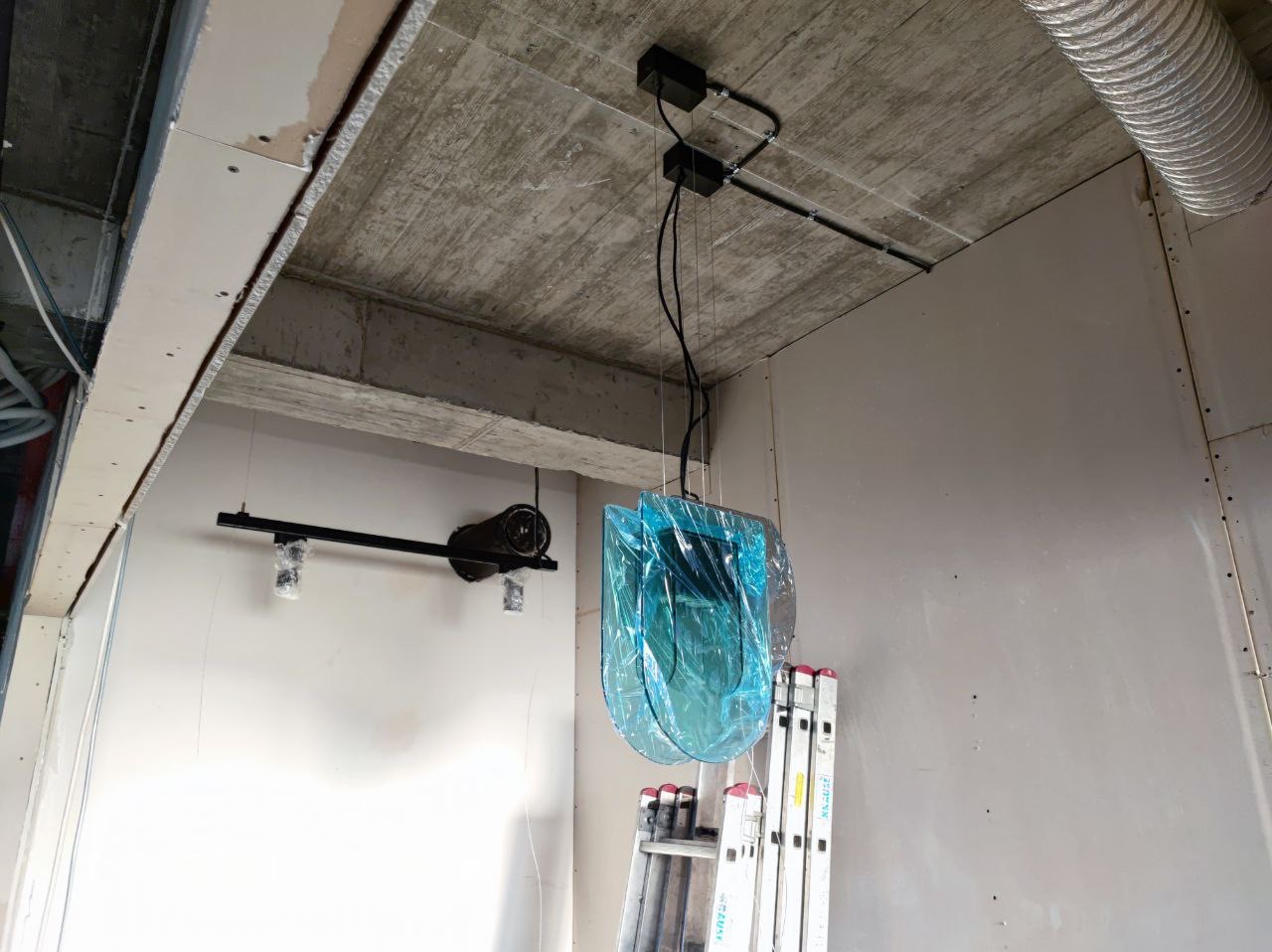 Montaż lampy wiszącej osłoniętej folią ochronną, widoczne przewody elektryczne pod sufitem, drabina i oświetlenie szynowe w surowym wnętrzu.