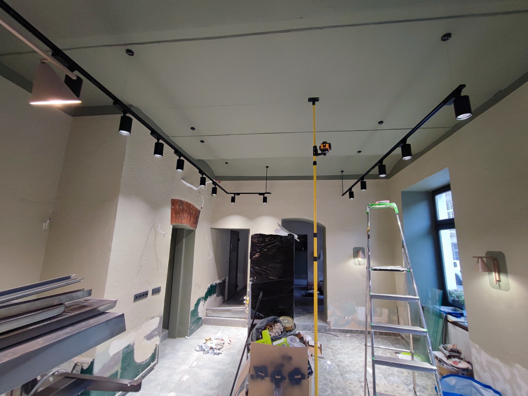 Wnętrze w trakcie remontu z zamontowanymi szynami oświetleniowymi z reflektorami, widoczny laser krzyżowy na wysięgniku, drabina, nieotynkowane ściany i materiały budowlane.