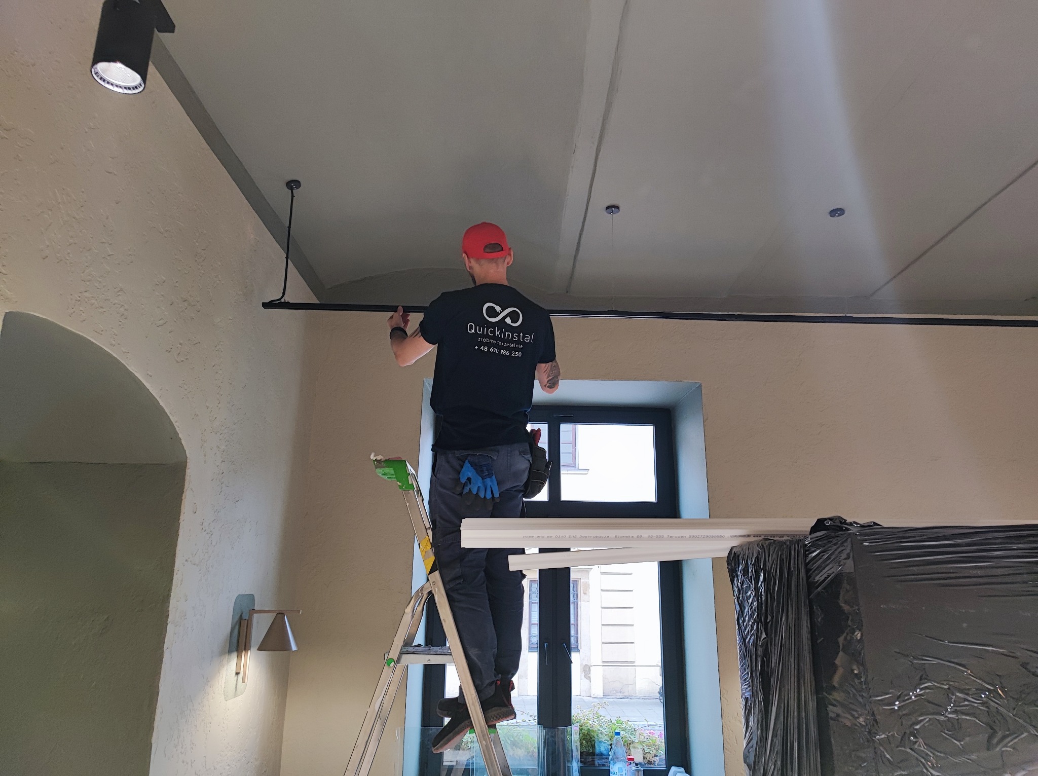 Instalator na drabinie montuje szynoprzewód oświetleniowy w pomieszczeniu z fakturą na ścianie i oknem w tle. Widoczne narzędzia w torbie na pasie i logo firmy na koszulce.
