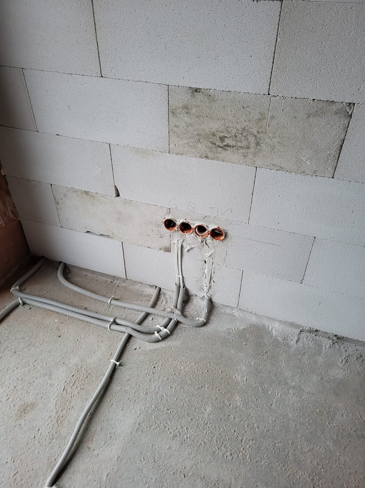 Ściana z betonu komórkowego z czterema otworami na przewody elektryczne, z których wychodzą szare rury karbowane, ułożone na surowym betonowym podłożu.