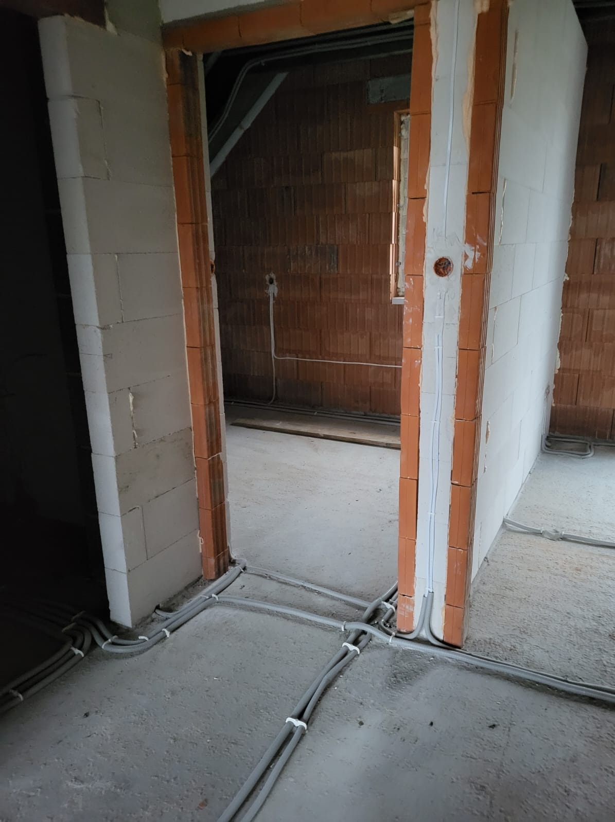 Szare kable elektryczne poprowadzone po surowej podłodze w nowo budowanym pomieszczeniu, widoczne ściany z cegły i pustaków, przygotowane otwory w ścianach i bruzdy pod instalacje.