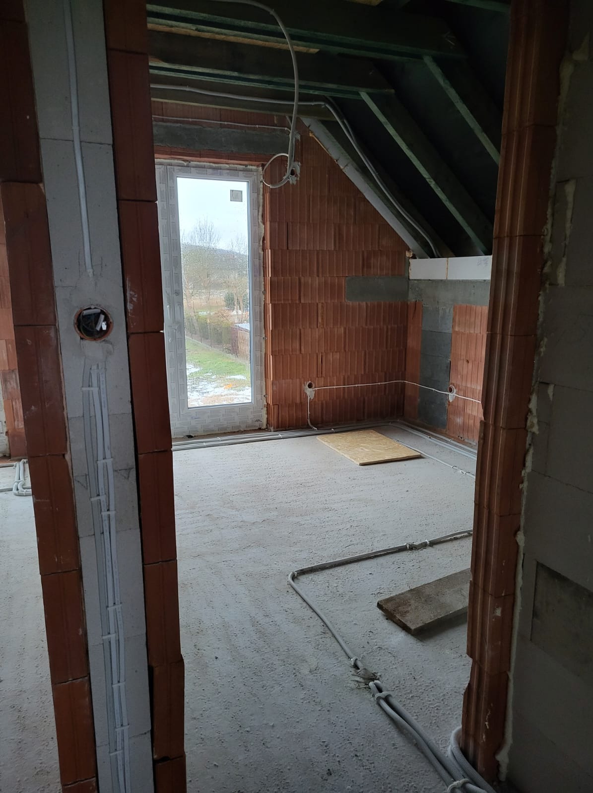 Surowy stan poddasza z widocznymi przewodami elektrycznymi ułożonymi wzdłuż ścian z czerwonej cegły i betonowej podłogi, okno z widokiem na zewnątrz, brak wykończenia.