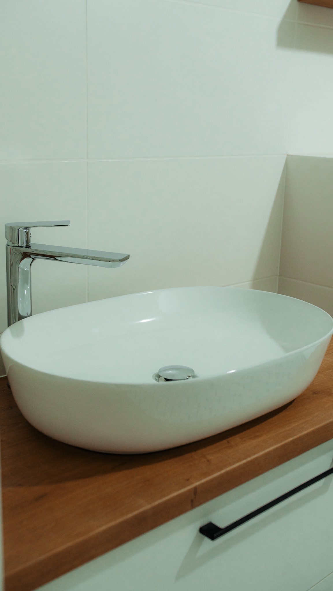 Nowoczesna umywalka nablatowa o owalnym kształcie z chromowaną baterią, osadzona na drewnianym blacie z minimalistycznymi czarnymi uchwytami szafki w tle białe płytki.