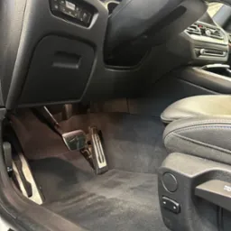Wnętrze samochodu z widocznymi pedałami, dywanikami i fragmentem skórzanego fotela z niebieską nicią, po czyszczeniu.