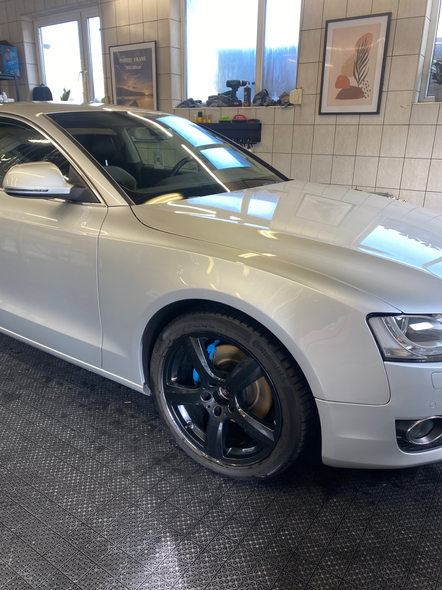 Srebrny samochód osobowy, widok z przodu na czarną felgę z niebieskimi zaciskami hamulcowymi, na podłodze z kratką, w tle okna i dekoracje na ścianie.