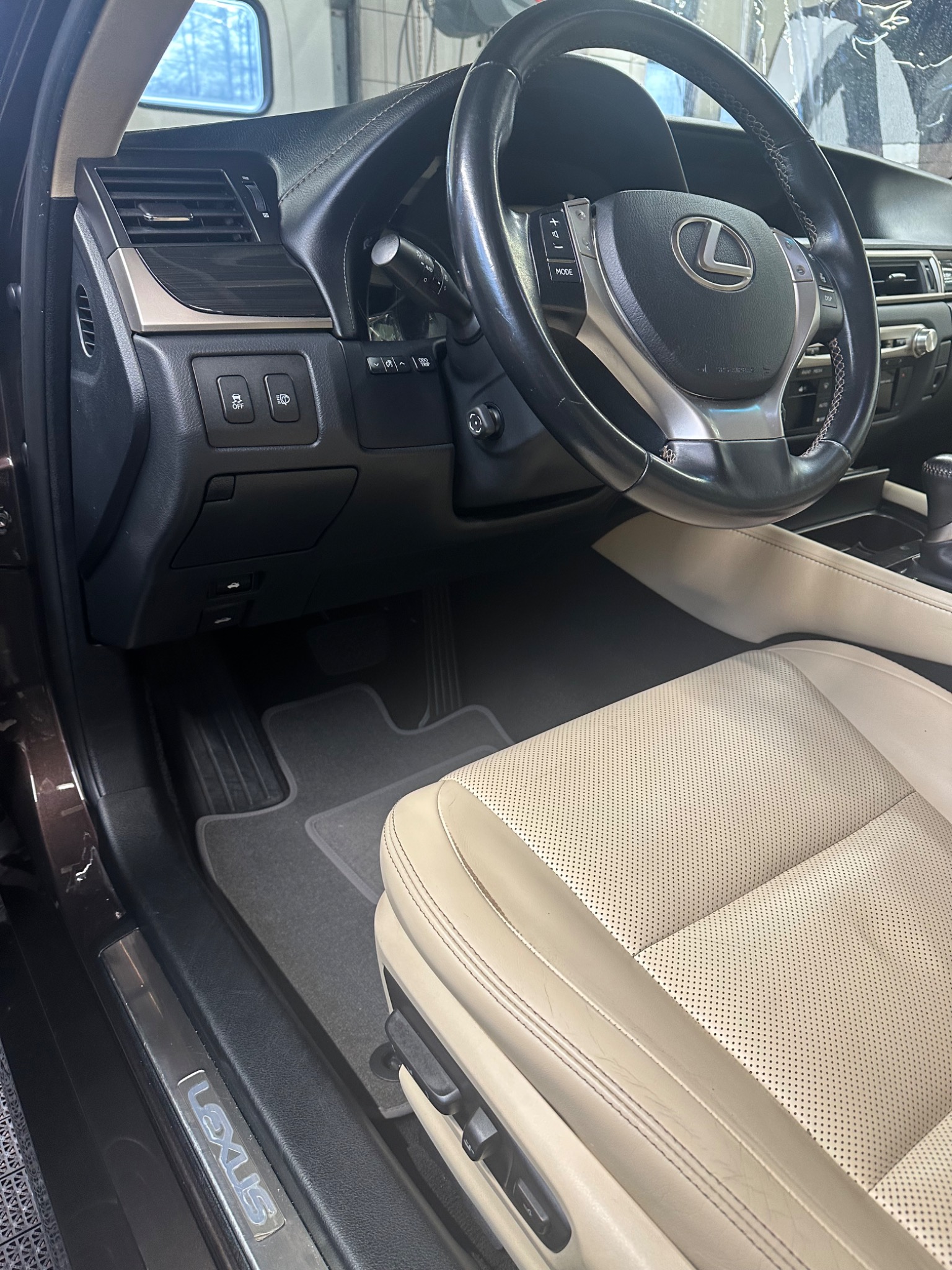 Wnętrze samochodu Lexus z beżową skórzaną tapicerką fotela kierowcy, widok na kierownicę z logo i deskę rozdzielczą.