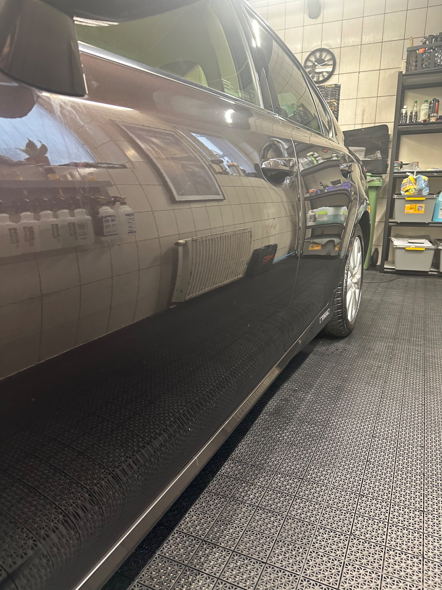 Błyszczący, ciemny samochód po umyciu, widoczny z boku na tle płytkowej ściany i podłogi w myjni, z refleksami otoczenia na karoserii.