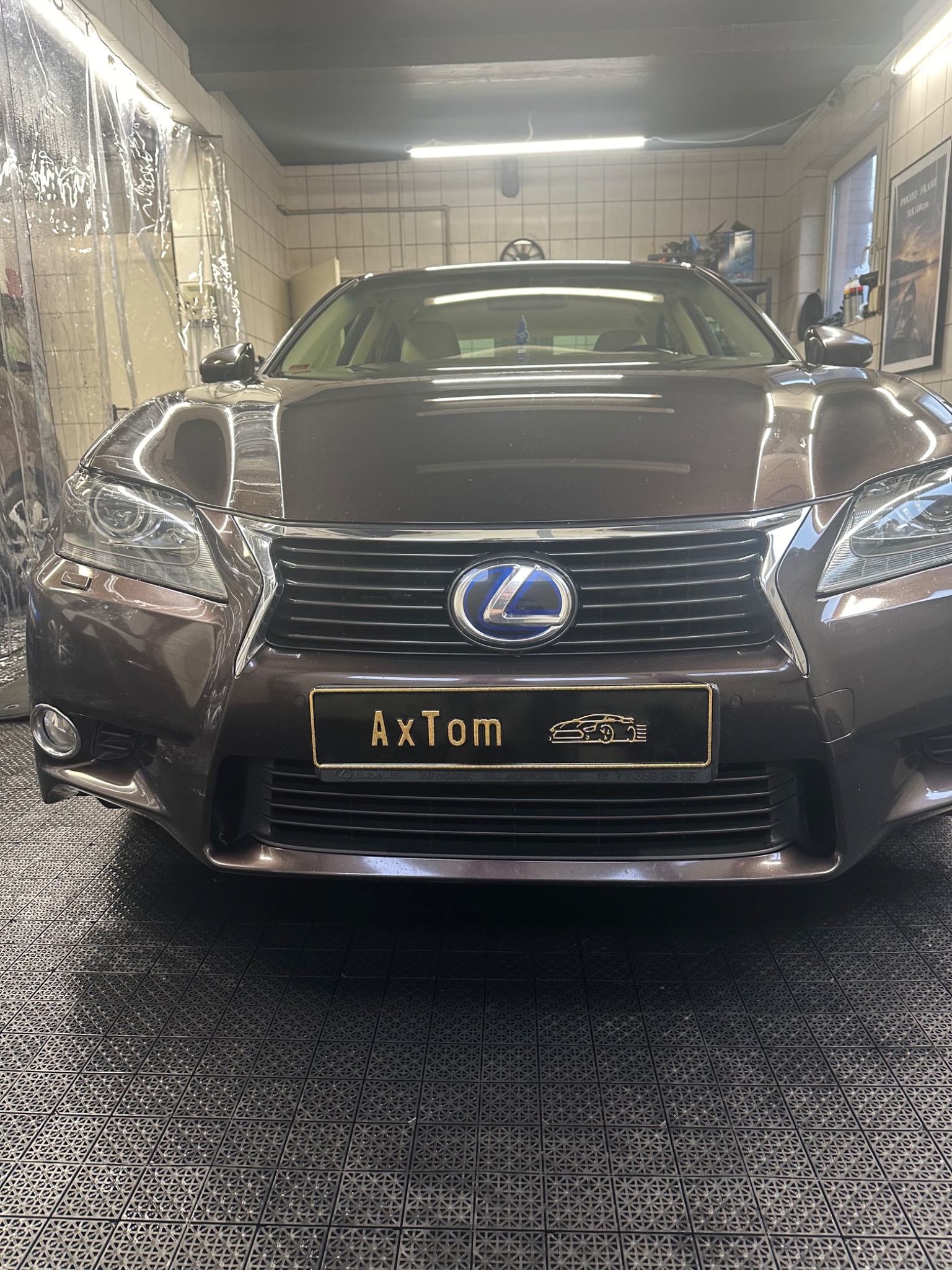 Brązowy Lexus GS z tablicą rejestracyjną 'AxTom' w myjni, widok z przodu na kratkę i logo