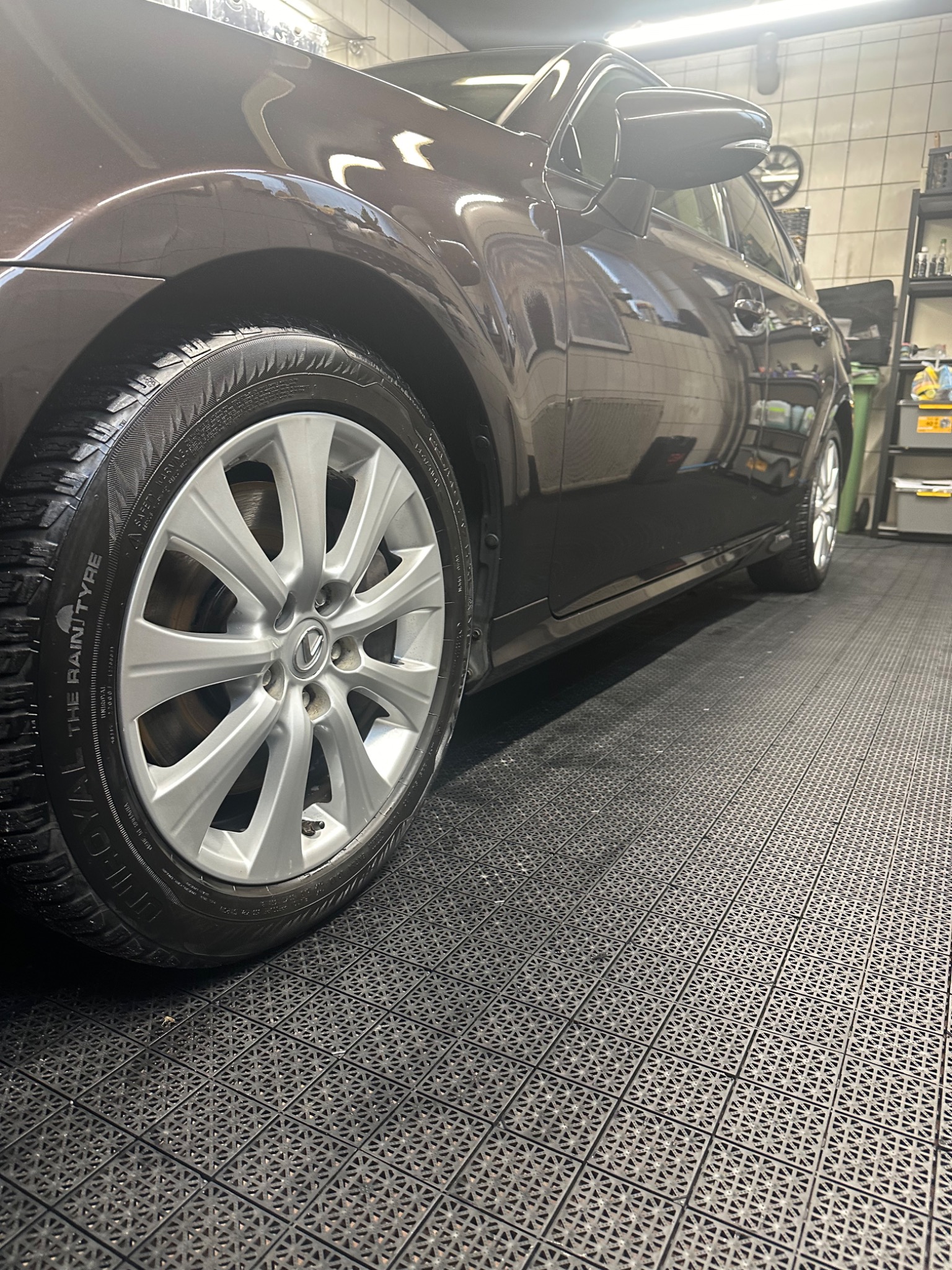 Ciemnobrązowy samochód Lexus po umyciu, widok z dołu na przednie koło i błyszczący lakier, na podłodze z gumowych płytek w myjni.