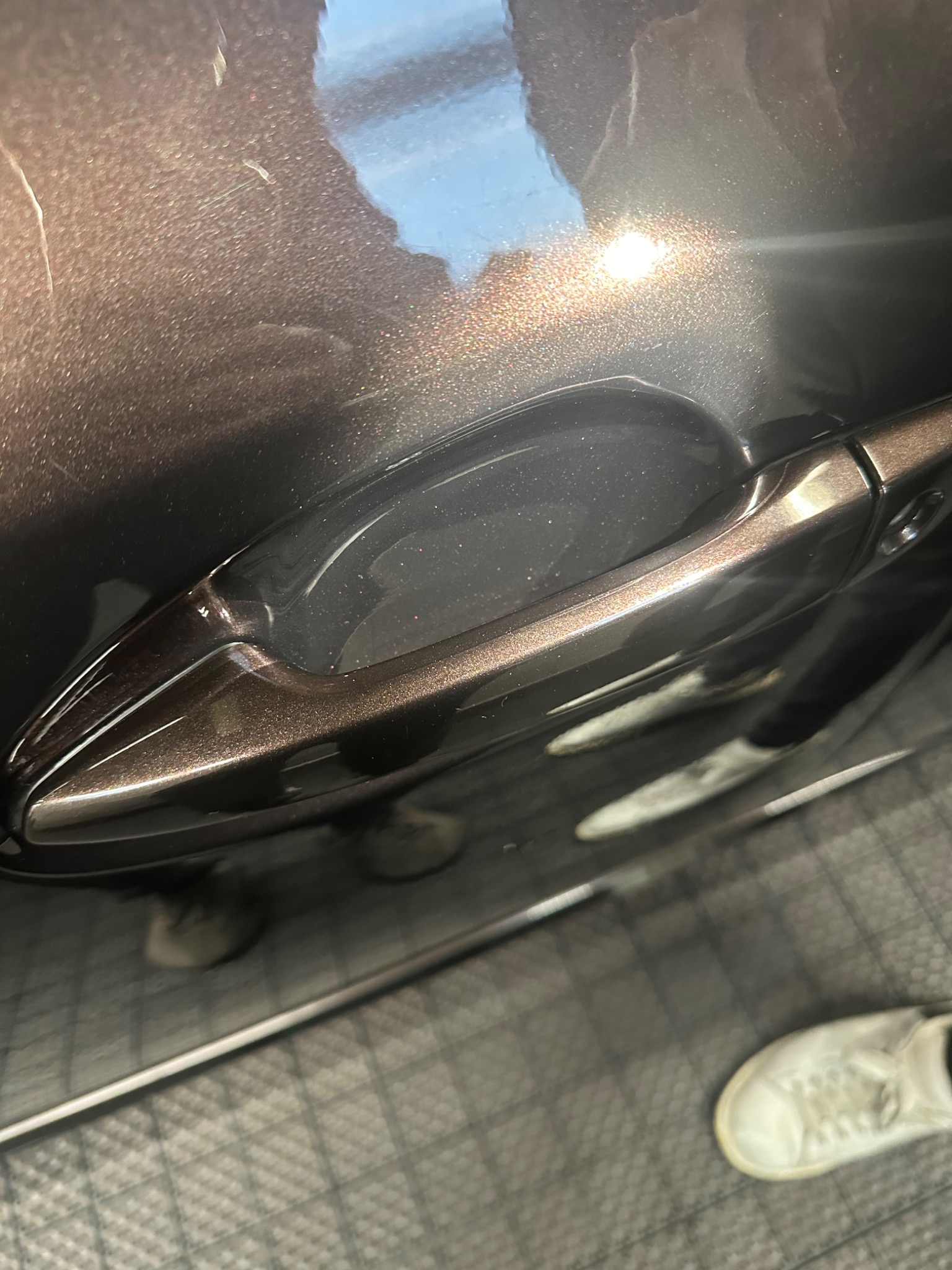 Błyszczący, brązowy lakier na samochodzie w okolicy klamki, widoczne drobne rysy i ślady po umyciu, odbicie światła słonecznego.